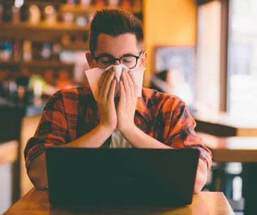 Man at a laptop blowing his nose, the allergies and asthma a damp home can bring on