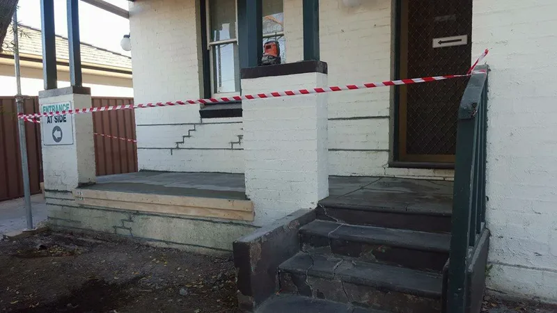 Summer Hill surgery verandah taped off during the repair, stepped cracks exposed in the stripped brickwork and a laser level set on the pier