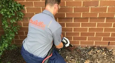 Technician kneeling at the garden bed beside the Lidcombe townhouse wall, feeding the GeoPoly™ injection hose into the ground