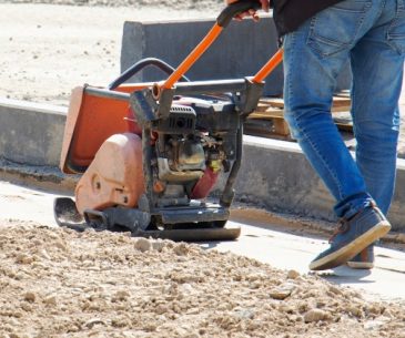 Worker compacting soil for foundation repair to prevent subsidence near concrete curb.