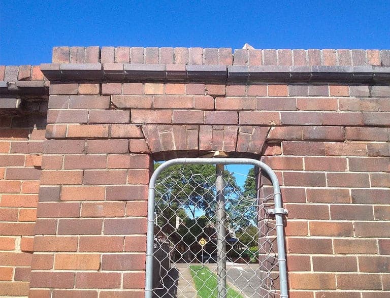 Brick wall with archway showing foundation cracks and subsidence near fence.