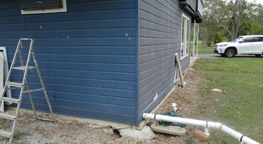 Blue house with foundation damage, wall cracks, exposed plumbing indicating subsidence repair need.