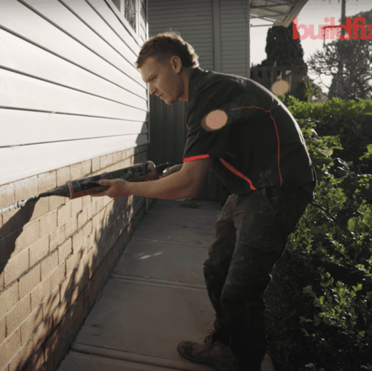Worker applying sealant to brick foundation gap for rising damp repair.