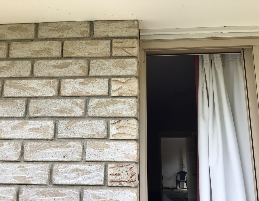 Light brown brick wall with early wall cracks indicating subsidence risk Sliding glass door beside brickwork showing subtle wall cracks and foundation issues Brick exterior displaying minor cracking, potential subsidence treatable with GeoPoly repair Cream-curtained entrance highlights early-stage wall cracks caused by foundation movement House exterior revealing masonry cracks, signs of subsidence suitable for structural repair.