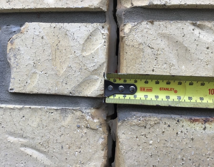 Yellow measuring tape measures brick wall crack indicating possible foundation subsidence repair.