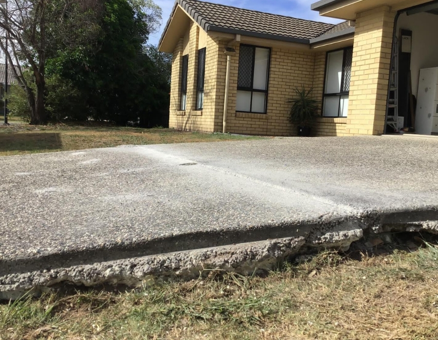 Concrete driveway subsidence near yellow brick house showing cracks and foundation issues.