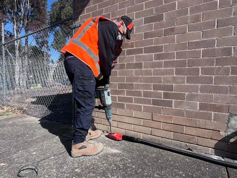 A worker in a safety vest and boots uses a power drill on the bottom section of a brick wall beside a chain-link fence outdoors.
