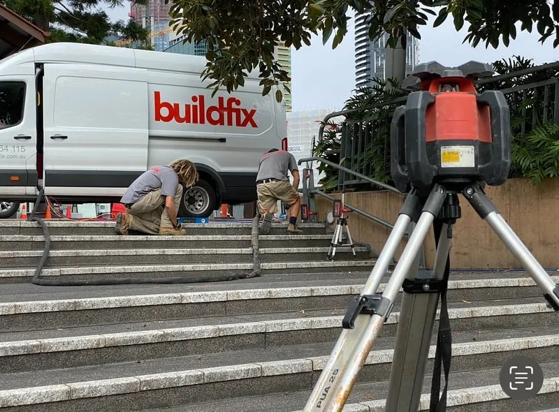Two workers repair stair railings near a Buildfix van, with construction equipment set up in the foreground.