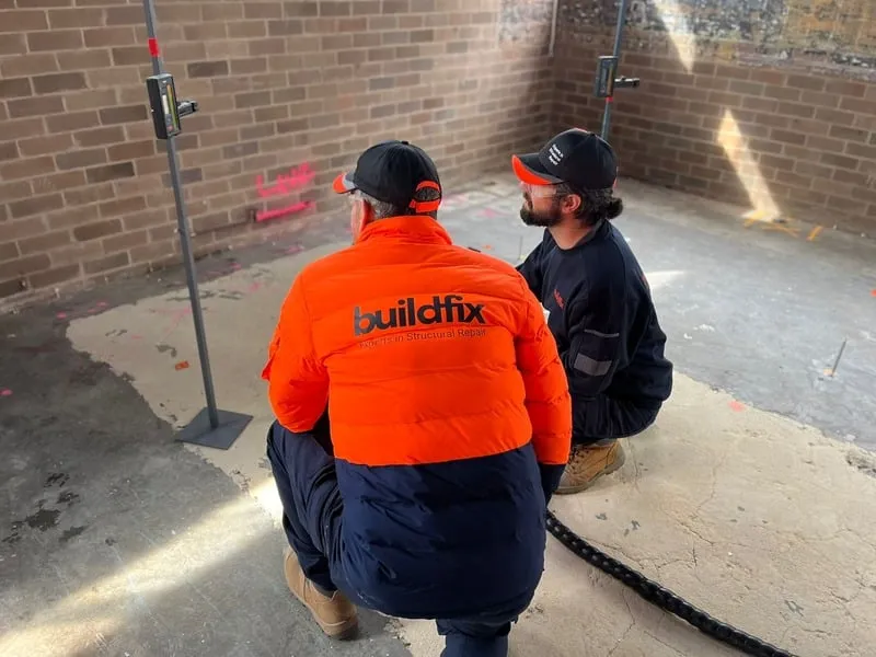 Two workers wearing buildfix uniforms kneel on a concrete floor inside a brick-walled room, observing equipment and red laser lines on the wall.